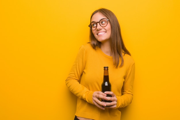 Young pretty caucasian woman smiling confident and crossing arms, looking up. She is holding a beer.