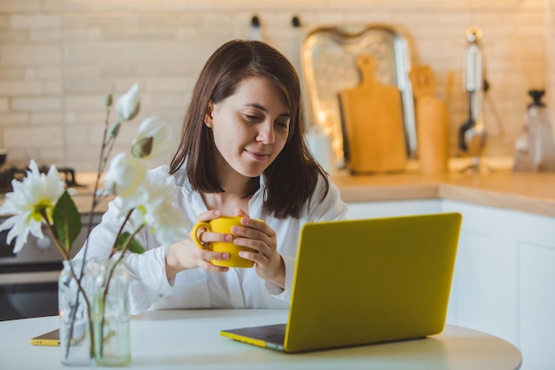 Young pretty caucasian woman talking on the phone sitting in front of laptop in the kitchen. business housewife. drinking coffee from yellow mug