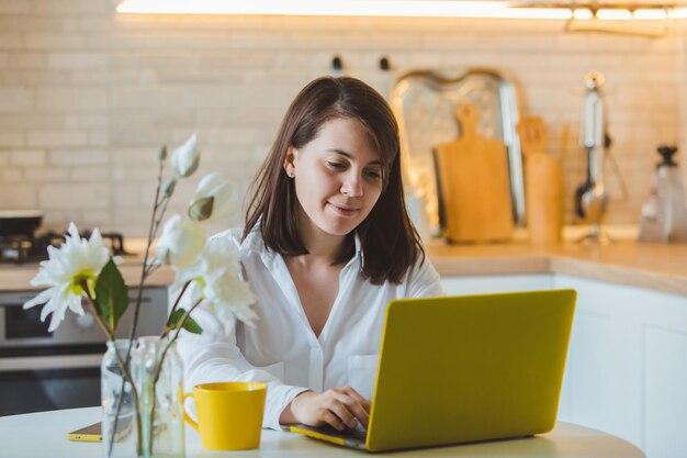 Young pretty caucasian woman talking on the phone sitting in front of laptop in the kitchen