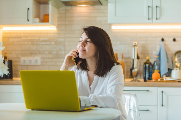 Young pretty caucasian woman talking on the phone sitting in front of laptop in the kitchen