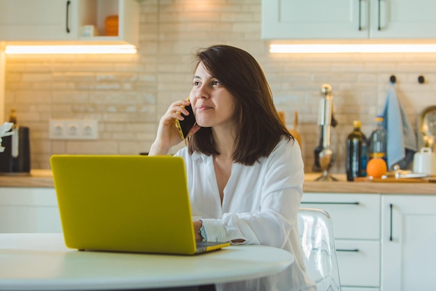 Young pretty caucasian woman talking on the phone sitting in front of laptop in the kitchen