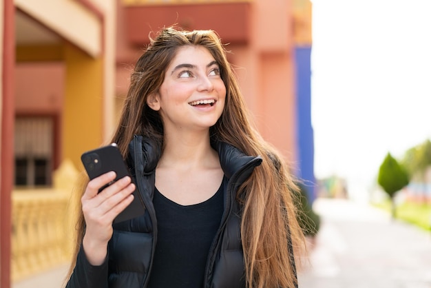 Young pretty caucasian woman using mobile phone at outdoors looking up while smiling