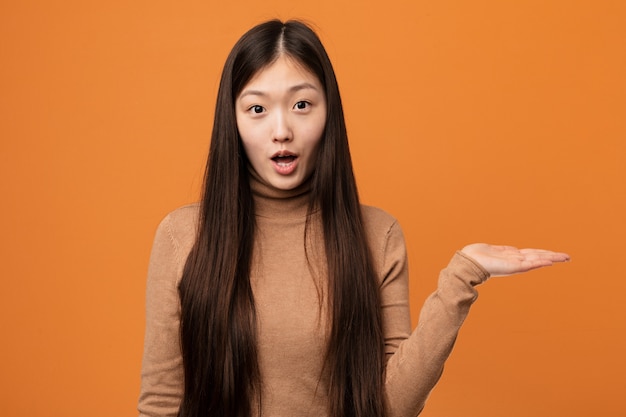 Young pretty chinese woman impressed holding copy space on palm.