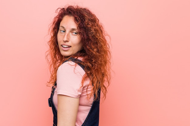 Photo young pretty ginger redhead woman wearing a jeans dungaree looks aside smiling, cheerful and pleasant.