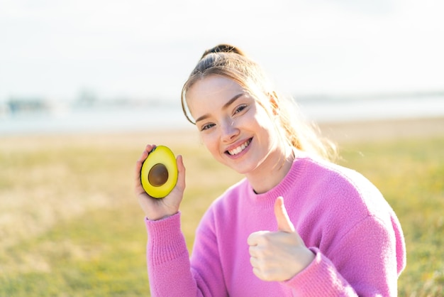 Young pretty girl holding an avocado at outdoors with thumbs up because something good has happened