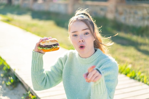 Young pretty girl holding a burger at outdoors surprised and pointing front