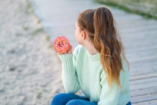 Young pretty girl holding a donut at outdoors in back position