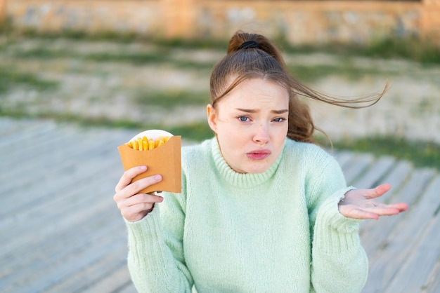 Young pretty girl holding fried chips at outdoors making doubts gesture while lifting the shoulders