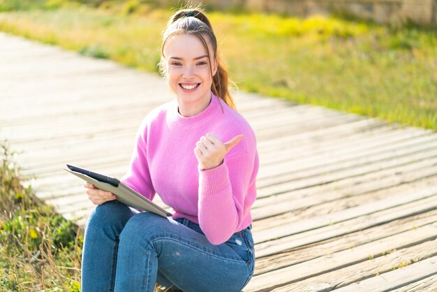 Young pretty girl holding a tablet at outdoors pointing to the side to present a product