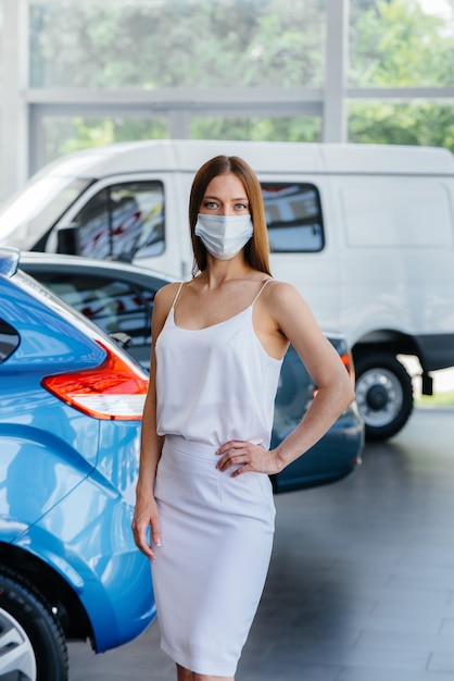 A young pretty girl inspects a new car at a car dealership in a mask during the pandemic. The sale and purchase of cars, in the period of pandemia.