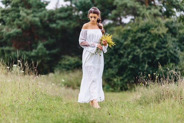 Young pretty girl with flower in light dressing walk at lawn near summer forest