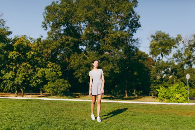 Young pretty girl with long brown hair dressed in light clothes staying on green lawn grass in the park on trees background. Summer sunny time.