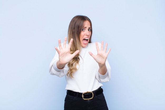 Young pretty hispanic woman feeling terrified, backing off and screaming in horror and panic, reacting to a nightmare against blue wall