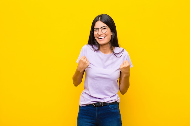 Young pretty latin woman shouting triumphantly, laughing and feeling happy and excited while celebrating success against flat wall