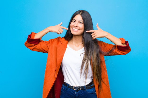 Young pretty latin woman smiling confidently pointing to own broad smile, positive, relaxed, satisfied attitude against flat wall