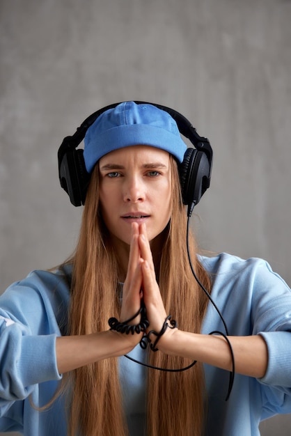 A young pretty longhaired dj girl in a blue sweater funny hat and headphones folded her hands in a p