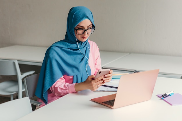 Young pretty modern muslim woman in hijab working on laptop in office room, education online