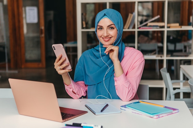 Young pretty modern muslim woman in hijab working on laptop in office room, education online