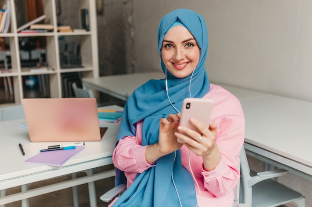 Young pretty modern muslim woman in hijab working on laptop in office room, education online