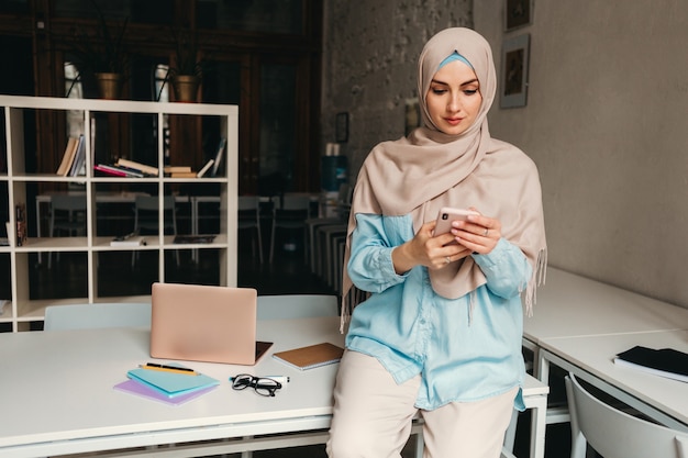 Young pretty modern muslim woman in hijab working in office room, education online