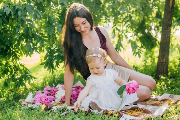 Young pretty mother with daughter lie on a plaid in the park. Family outdoor recreation. Family summer picnic in the park. Mother and daughter outdoors surrounded by pink peonies.