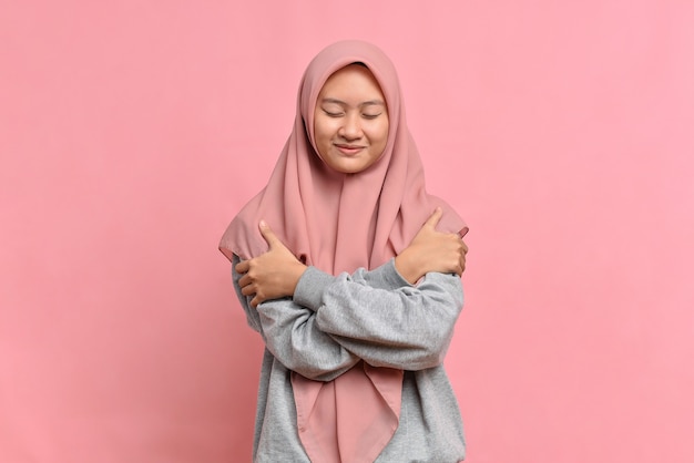 Young pretty muslim woman feeling in love, smiling, cuddling and hugging self, staying single and egocentric against pink background.