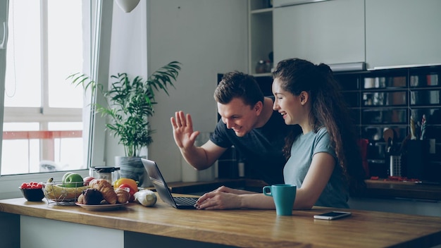 Young pretty positive caucasian couple sitting at table and have video call on laptop computer at home