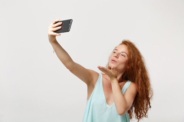 Young pretty redhead woman girl in casual light clothes posing isolated on white background. People lifestyle concept. Mock up copy space. Doing selfie shot on mobile phone, blowing sending air kiss.