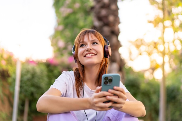 Young pretty redhead woman at outdoors listening music with the mobile and happy