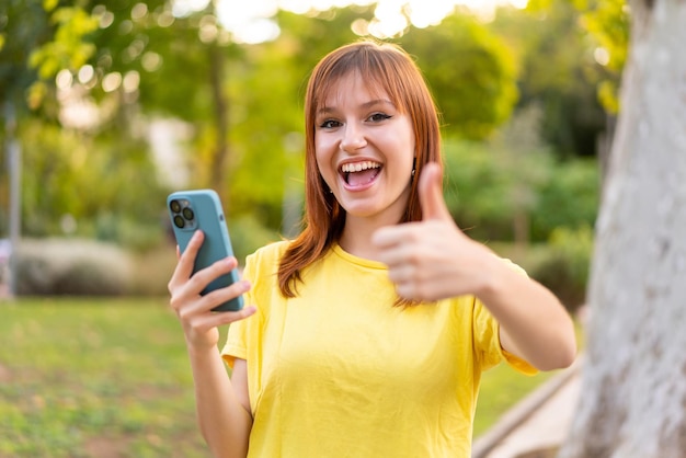 Young pretty redhead woman at outdoors using mobile phone while doing thumbs up