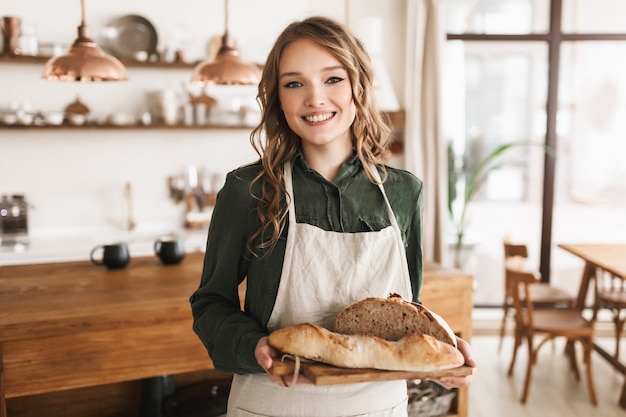 Young pretty smiling woman with wavy hair in white apron holding bread on board in hands, happily 