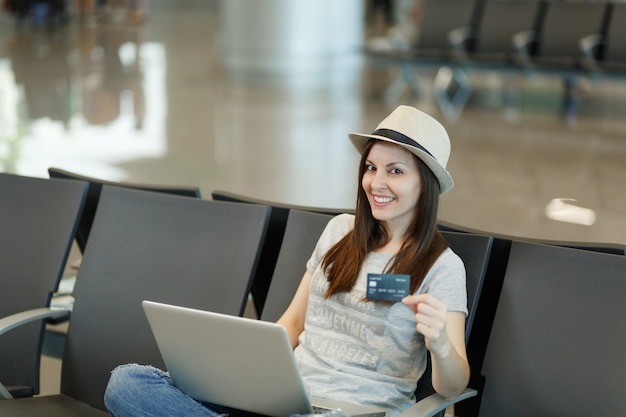 Young pretty traveler tourist woman in hat sitting, working on laptop, holding credit card while waiting in lobby hall at airport