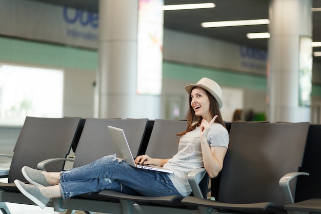 Young pretty traveler tourist woman working on laptop holding finger up having idea waiting in lobby hall at international airport