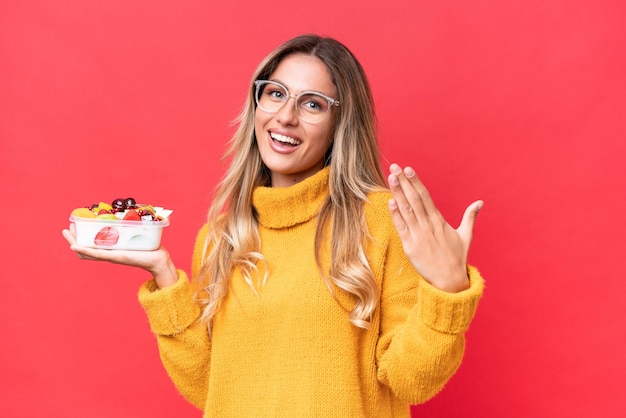 Photo young pretty uruguayan woman holding a bowl of fruit isolated on red background inviting to come with hand happy that you came