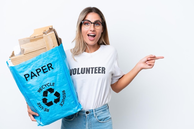 Young pretty Uruguayan woman holding a recycling bag full of paper to recycle isolated on white chroma background surprised and pointing finger to the side