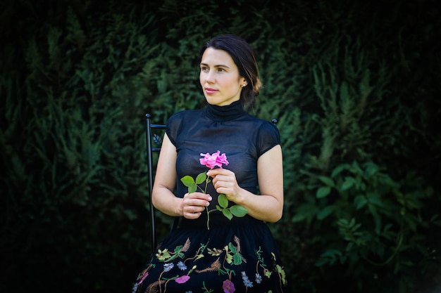 Young pretty woman in black clothes sits on a chair in the garden near a bush with a rose in her hands