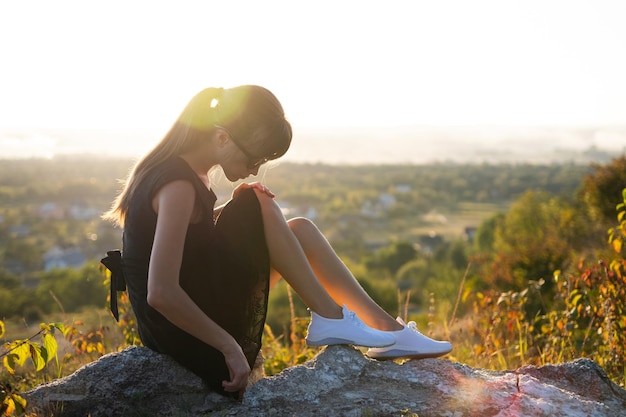 Young pretty woman in black short summer dress sitting on a rock relaxing outdoors at sunset Fashionable female enjoying warm evening in nature