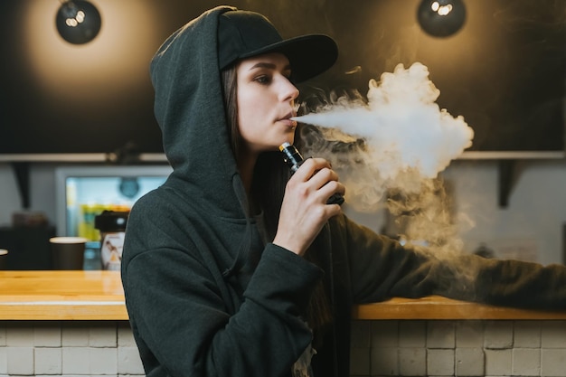 Young pretty woman in cap smoke an electronic cigarette at the vape shop Hiphop style Closeup