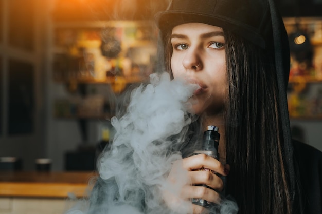 Young pretty woman in cap smoke an electronic cigarette at the vape shop Hiphop style Closeup