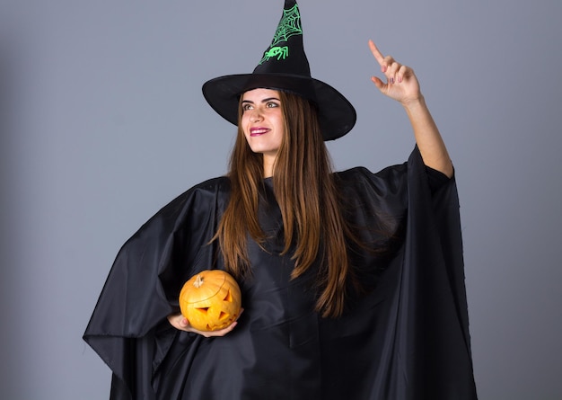 Young pretty woman in costume of witch with black hat holding a pumpkin on blue background in studio