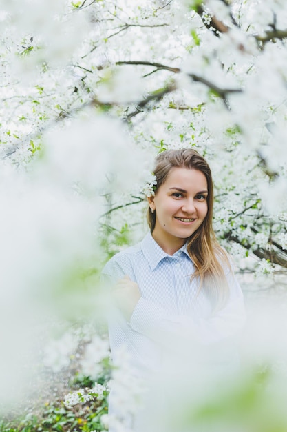 Young pretty woman enjoys standing near flowering spring tree A girl wearing beige hat and white dress smiles among blooming apple trees Spring season concept