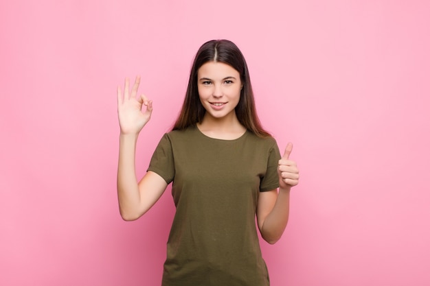 Young pretty woman feeling happy, amazed, satisfied and surprised, showing okay and thumbs up gestures, smiling against pink wall