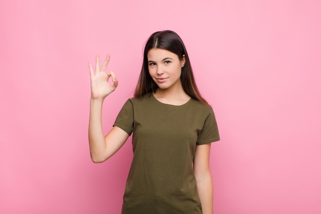 Young pretty woman feeling happy, relaxed and satisfied, showing approval with okay gesture, smiling against pink wall