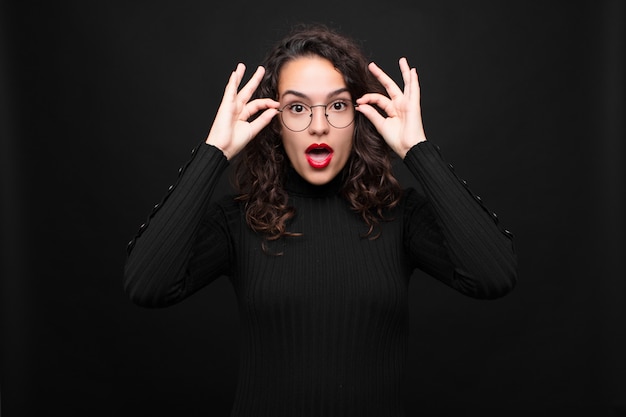 Photo young pretty woman feeling shocked, amazed and surprised, holding glasses with astonished, disbelieving look against black wall.