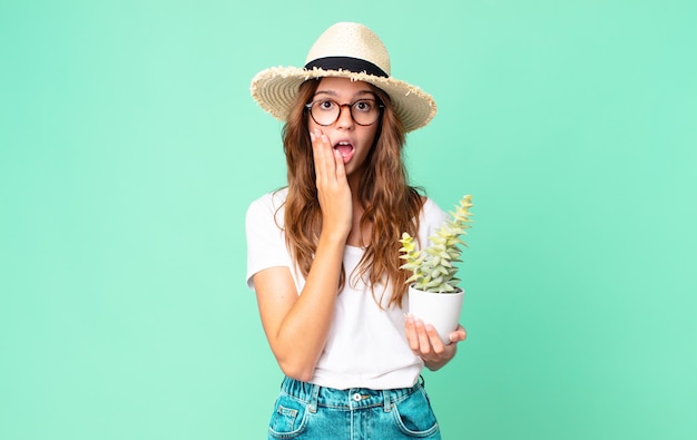 Young pretty woman feeling shocked and scared with a straw hat and holding a cactus