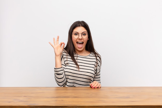 Young pretty woman feeling successful and satisfied, smiling with mouth wide open, making okay sign with hand sitting at a table