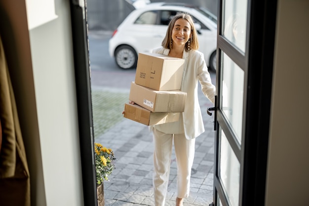 Young pretty woman going home, carrying a pile of parcels, coming home by a city car. View through the house door