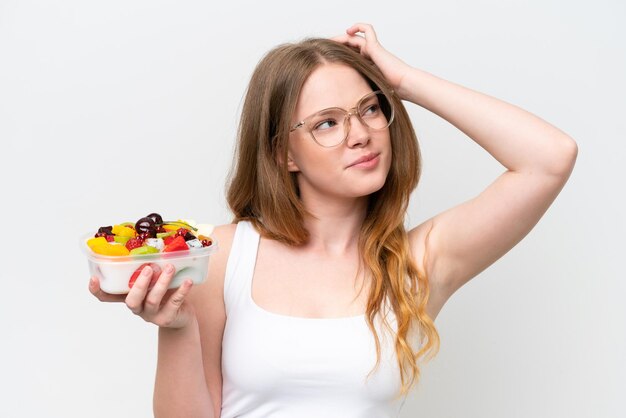 Young pretty woman holding a bowl of fruit isolated on white background having doubts and with confuse face expression