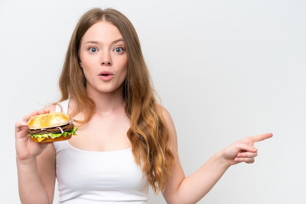 Young pretty woman holding a burger isolated on white background surprised and pointing side