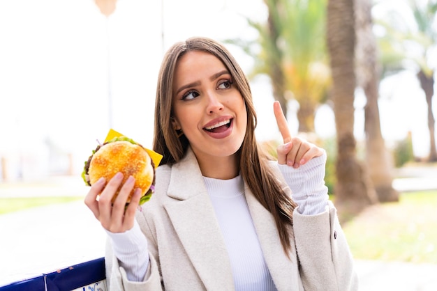 Young pretty woman holding a burger at outdoors intending to realizes the solution while lifting a finger up